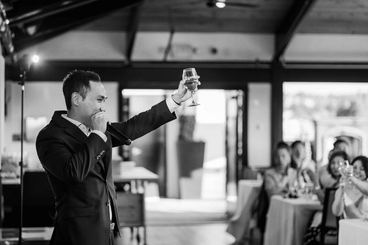 Best man raises glass during toast, facing the audience as they clap