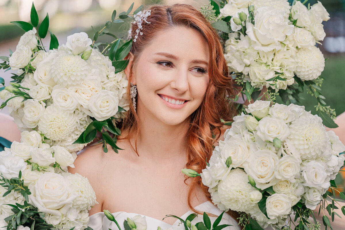 A smiling redheaded bride surrounded by white and green flowers on her wedding day in Cary enjoying her Raleigh wedding photography session by JoLynn Photography
