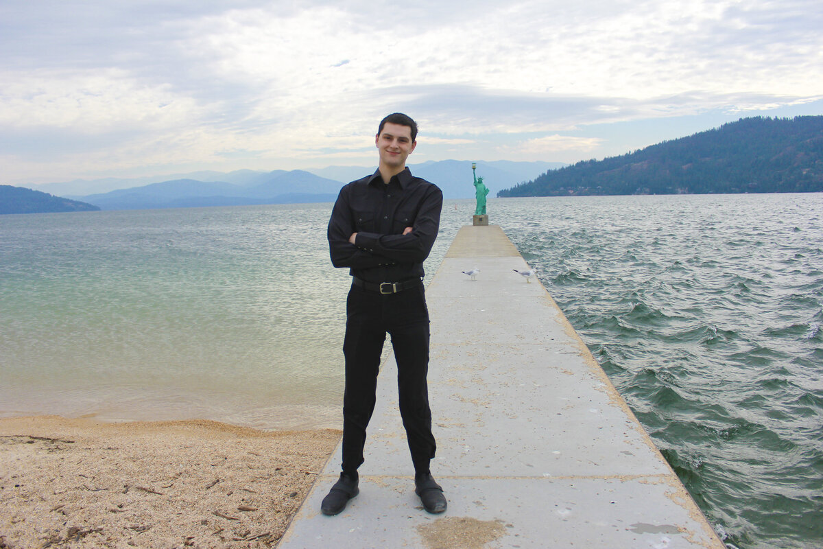 photo of a Man standing on a pier  near lake with a statue of liberty in background