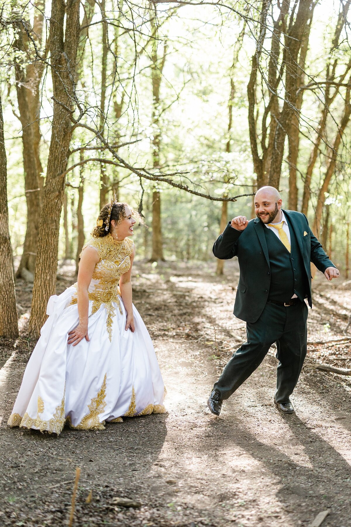 A portrait of a bride laughing while the groom is dancing playfully in a wooded setting after their backyard wedding ceremony in Fort Worth, Texas. The bride is on the left is a wearing an elaborately decorated white and gold dress with a golden sun crown. The groom is on the right and is wearing a dark green suit with a yellow tie and pocket square.