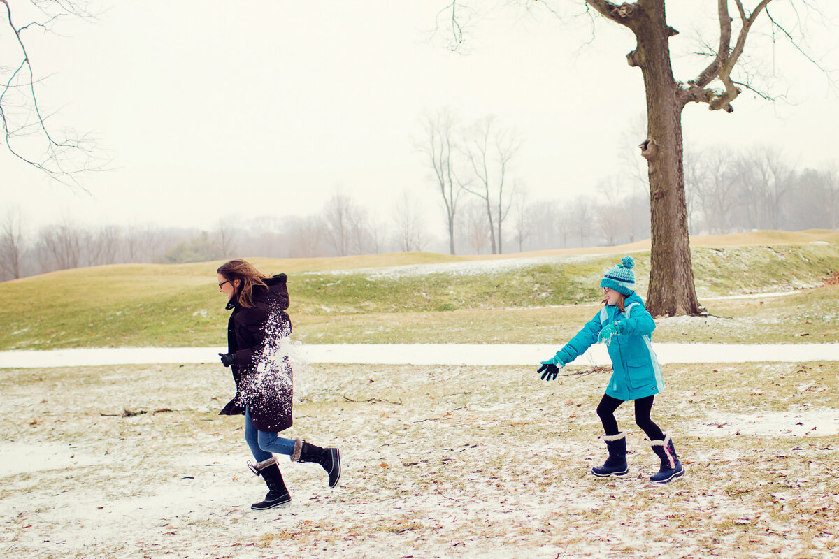 Girl in blue coat and blue hat throws a snowball and hits her mom wearing a black coat as she runs away.