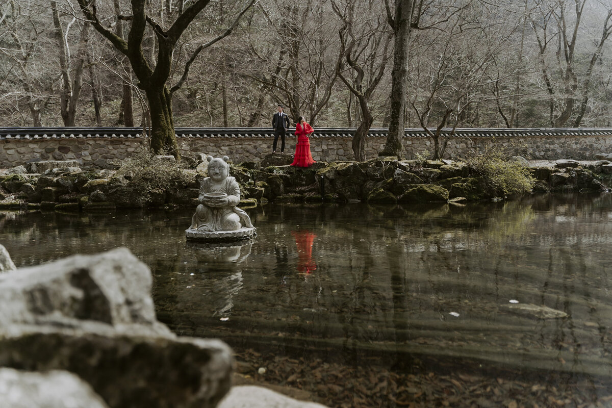 bride wearing a red dress and groom wearing a black suit looking at the trees during their pre-wedding photoshoot in Korea