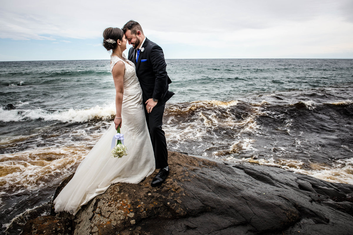 Bride and groom kiss on a rock in front of crashing waves of Lake Superior.