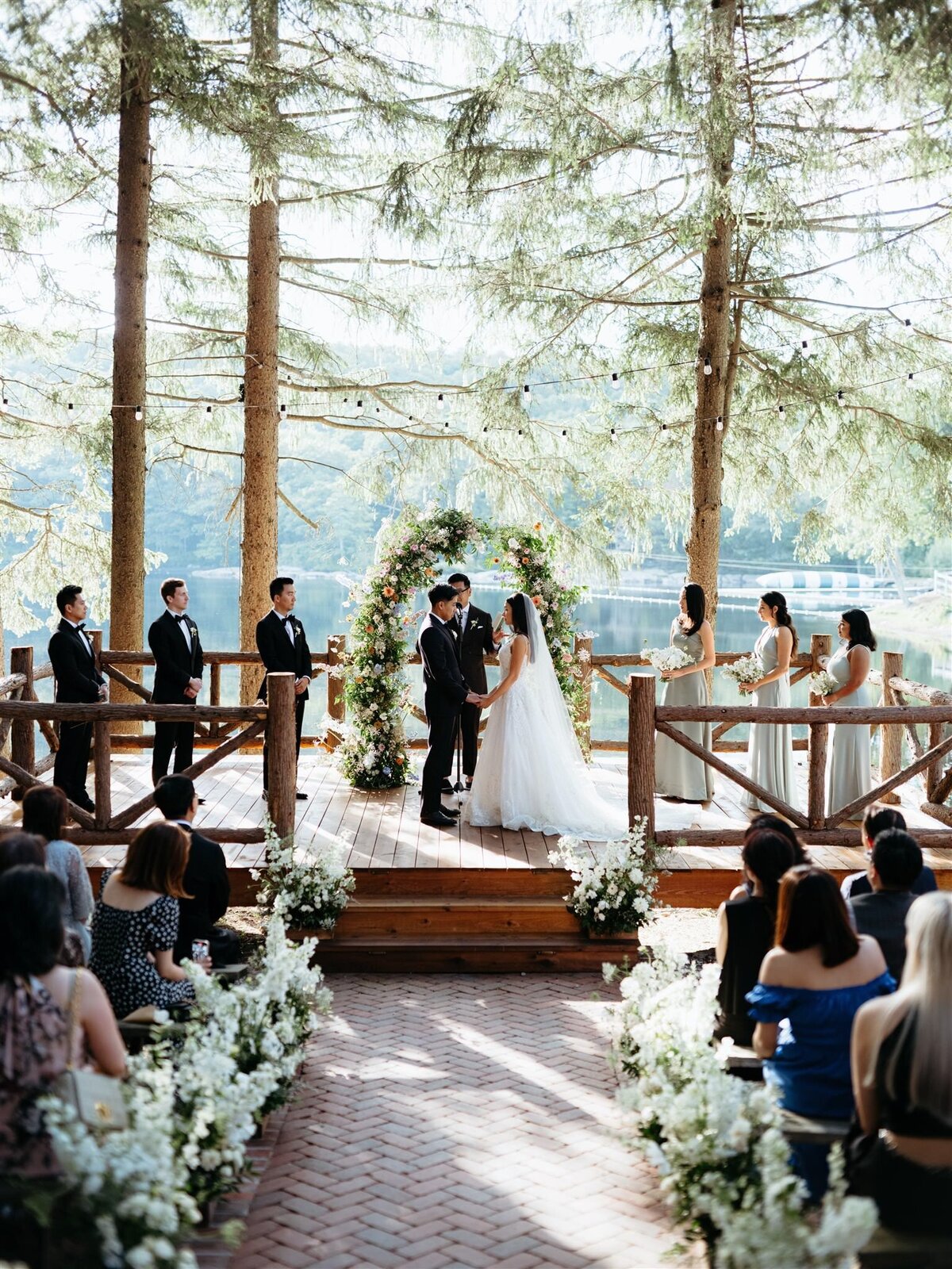 Bride and groom embrace for kiss during wedding ceremony on deck in front of stunning floral arbor, pine trees, and lake in Hudson Valley.