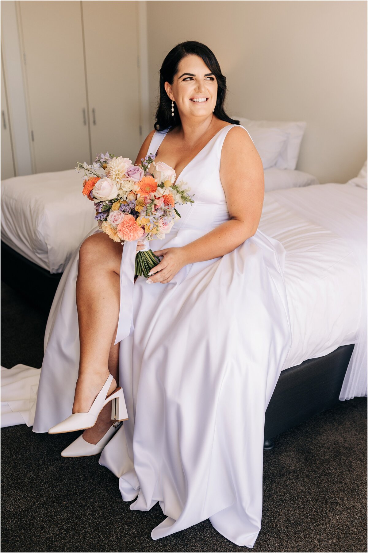 bride sitting on white bed in hotel christchurch clearwater resort with bouquet orange and pink, smiling with white shoes from mi piaci