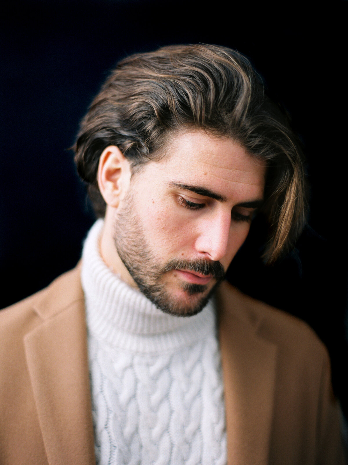 Man looks downward in camel coat and white turtleneck sweater