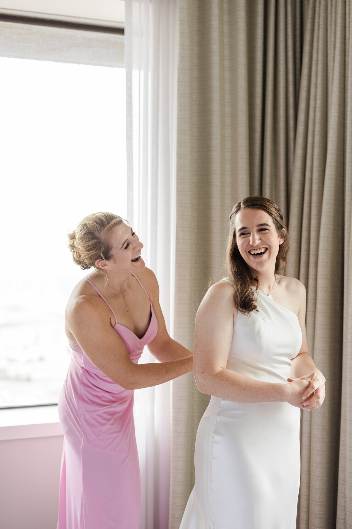 A bride being helped into her dress by one of her bridesmaids while getting ready at The Westin Dallas Downtown in Dallas, Texas before her wedding ceremony. The bride is wearing a simple but elegant white dress, and her bridesmaid is wearing a pink dress. They are both laughing joyfully.