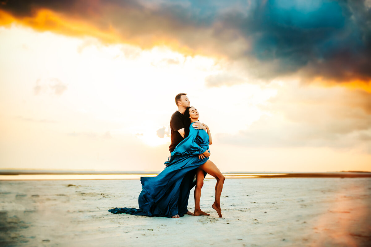 incredible colorful moody beach couples portrait with a eomen wearing a couture blue flow dress blowing in the wind