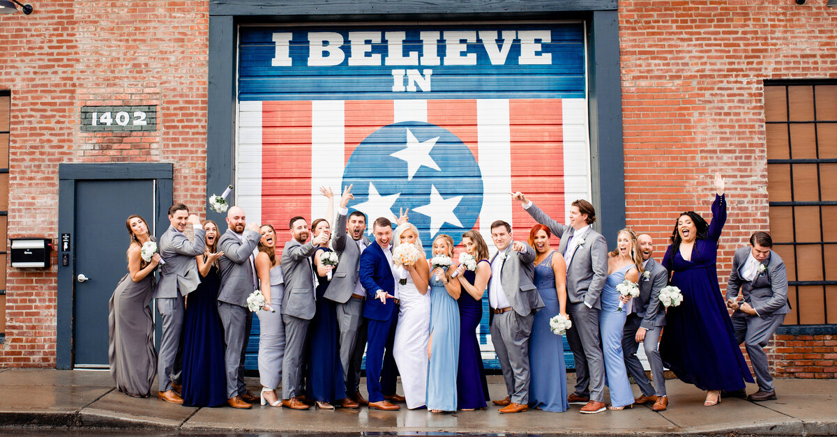 Wedding party wearing shades of blue and grey dancing together in front of the I believe in Nashville wall near Marathon Village