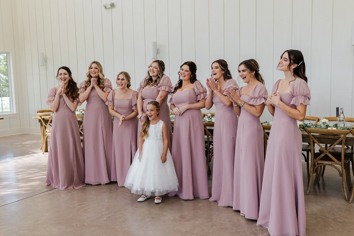 Candid shot of a group of bridesmaids and a flower girl all reacting joyfully to seeing the bride (out of the shot) for the first time at a wedding in Dallas, Texas. The bridesmaids are all wearing lavender dresses, and the flower girl is wearing a sleeveless white dress.