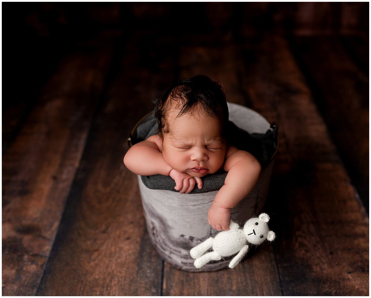 A baby  in a bucket with chin on one hand and the other hand out of the bucket holding a white teddy bear.