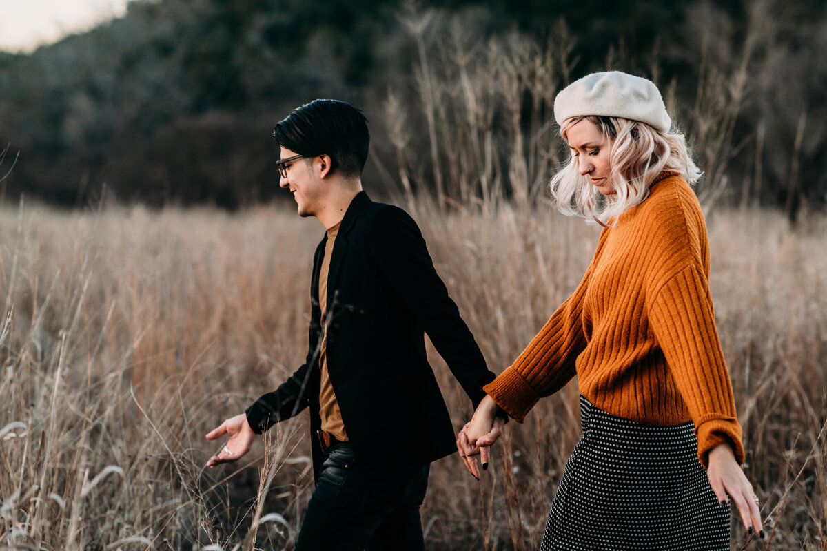 Couples Photography, Man in a blazer walks through the field holding a woman's hand in a sweater walk through tall dry grass