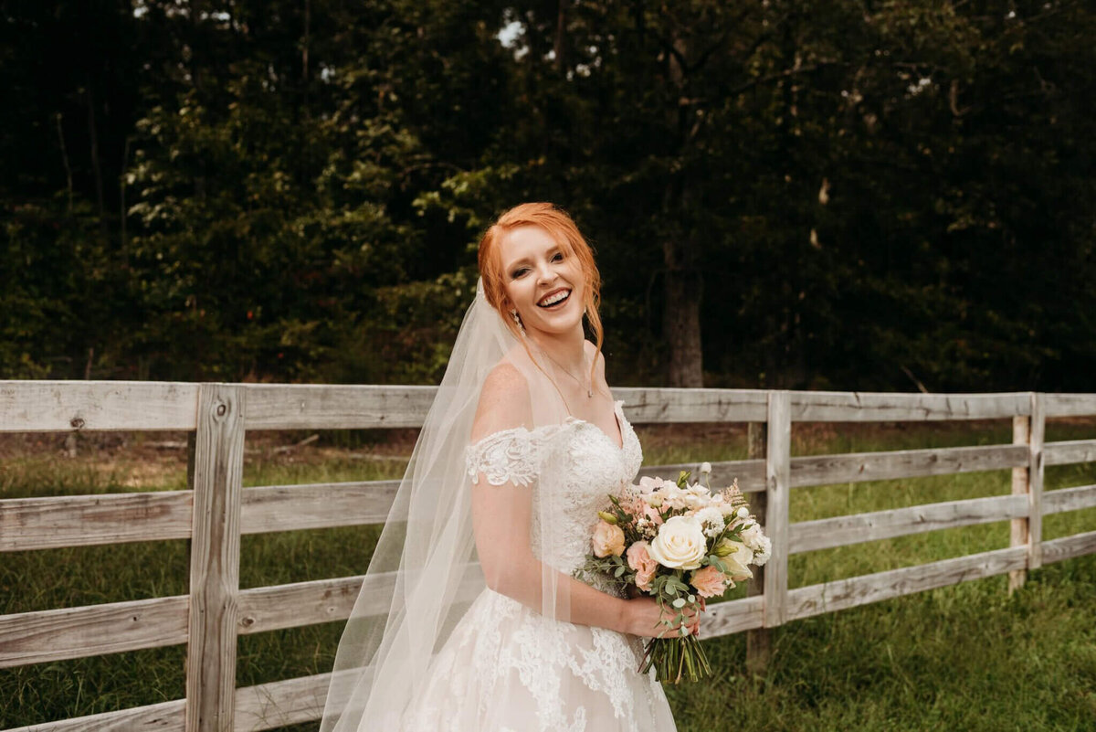 Photo of a bride smiling in front of a wooden fence while holding a small bouquet and wearing a veil