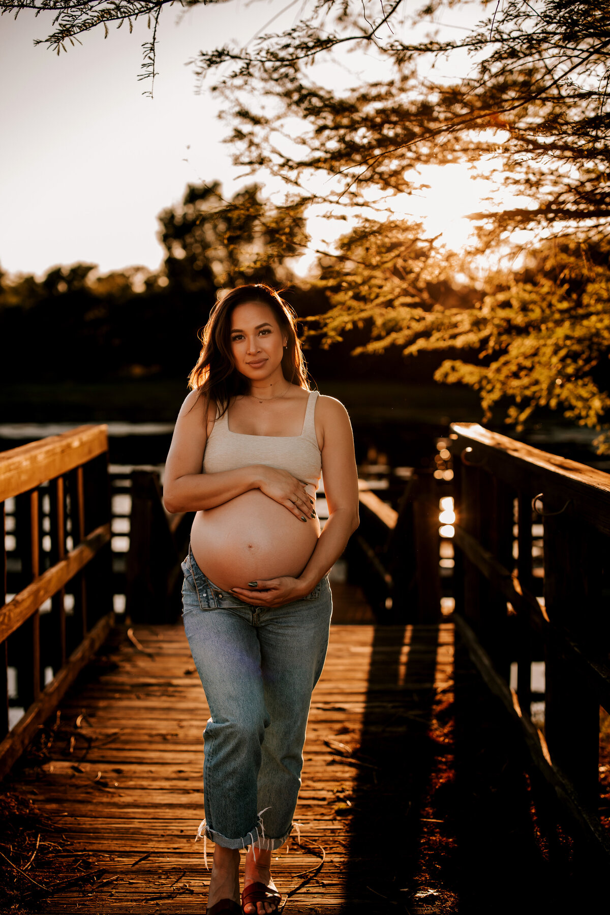 pregnant mom stands on boat dock during glden hour while wearing a tan bralette and jeans while holding bare belly