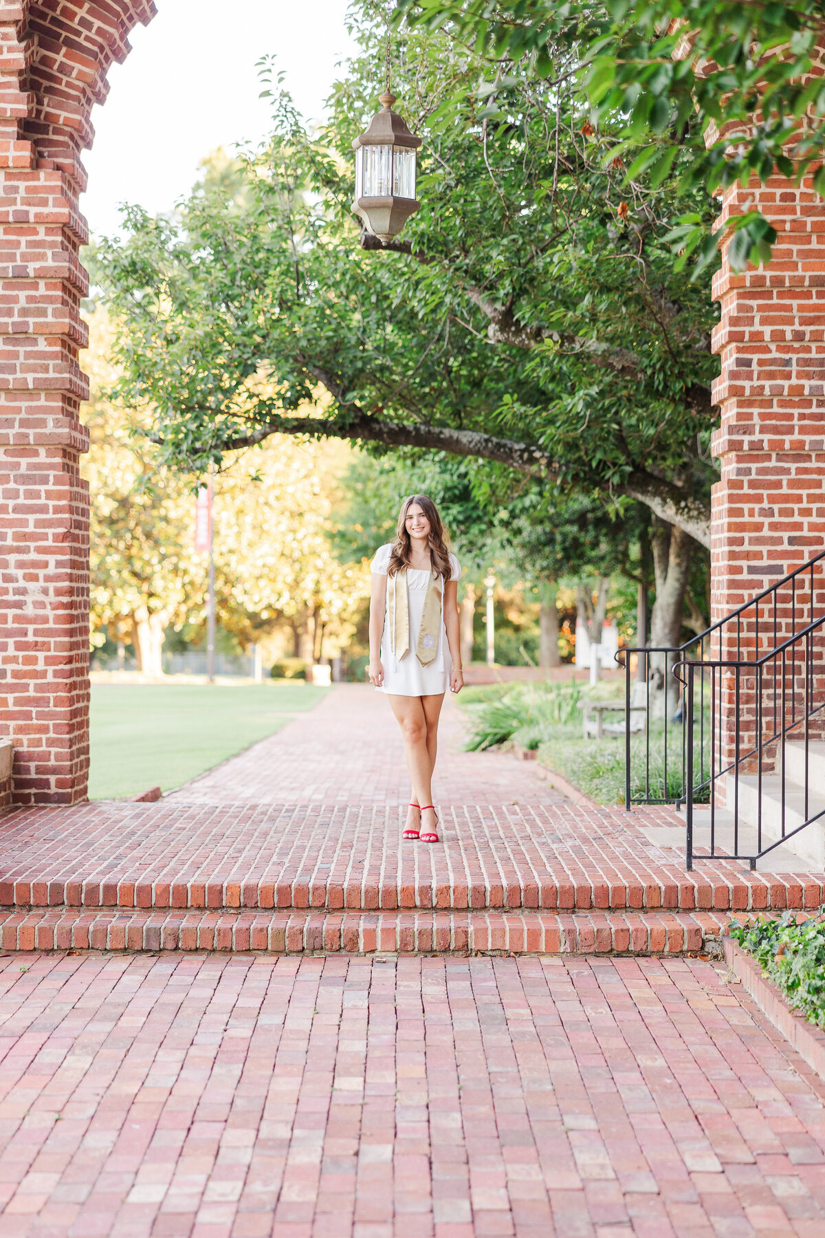 A person stands confidently on a brick pathway between archways, surrounded by lush greenery and trees. The individual wears a light-colored dress and red shoes, hands on hips. A hanging lantern is visible above, perfectly capturing the essence of a senior portrait by a talented photographer in Fayetteville, NC.