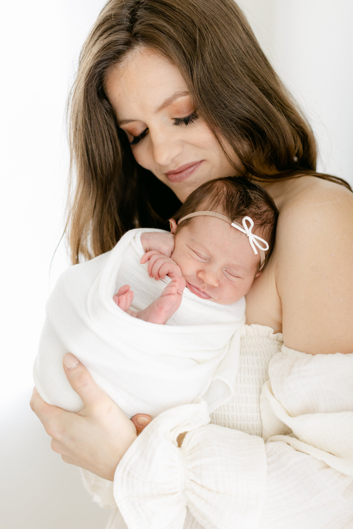 brunetter mom holding her baby girl swaddled in a white blanket in a bright and airy studio photographed by Philadelphia Newborn Photographer Tara Federico