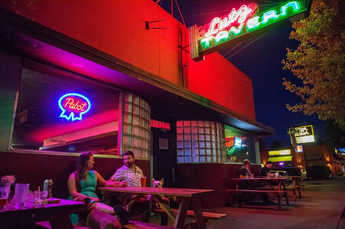 a hip couple and their dog sits at a picnic table underneath the neon lit signs of The Lutz Tavern in Woodstock at night