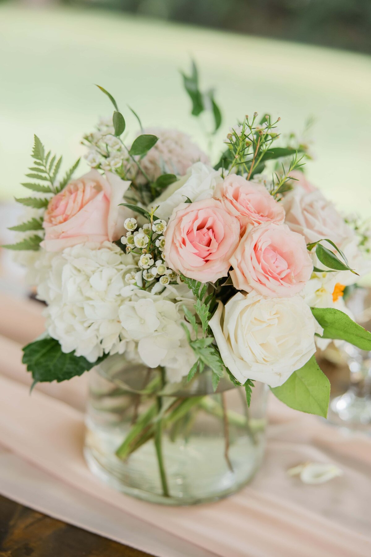A floral arrangement in a glass vase featuring pink roses, white hydrangeas, and green foliage on a table with a pink runner, perfect for park farm winery weddings.