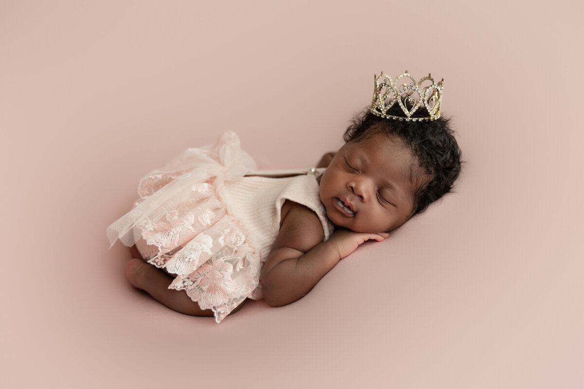A newborn baby in a pink dress on her belly wearing a tiny crown