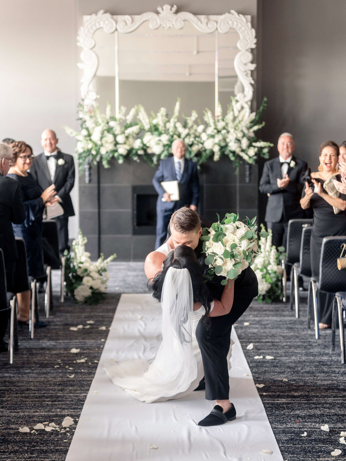 A newlywed couple shares a kiss on a white aisle. The groom holds the bride in a romantic dip. Guests in formal attire applaud. The backdrop features a large mirror and elaborate floral arrangements in a bright, elegant setting.