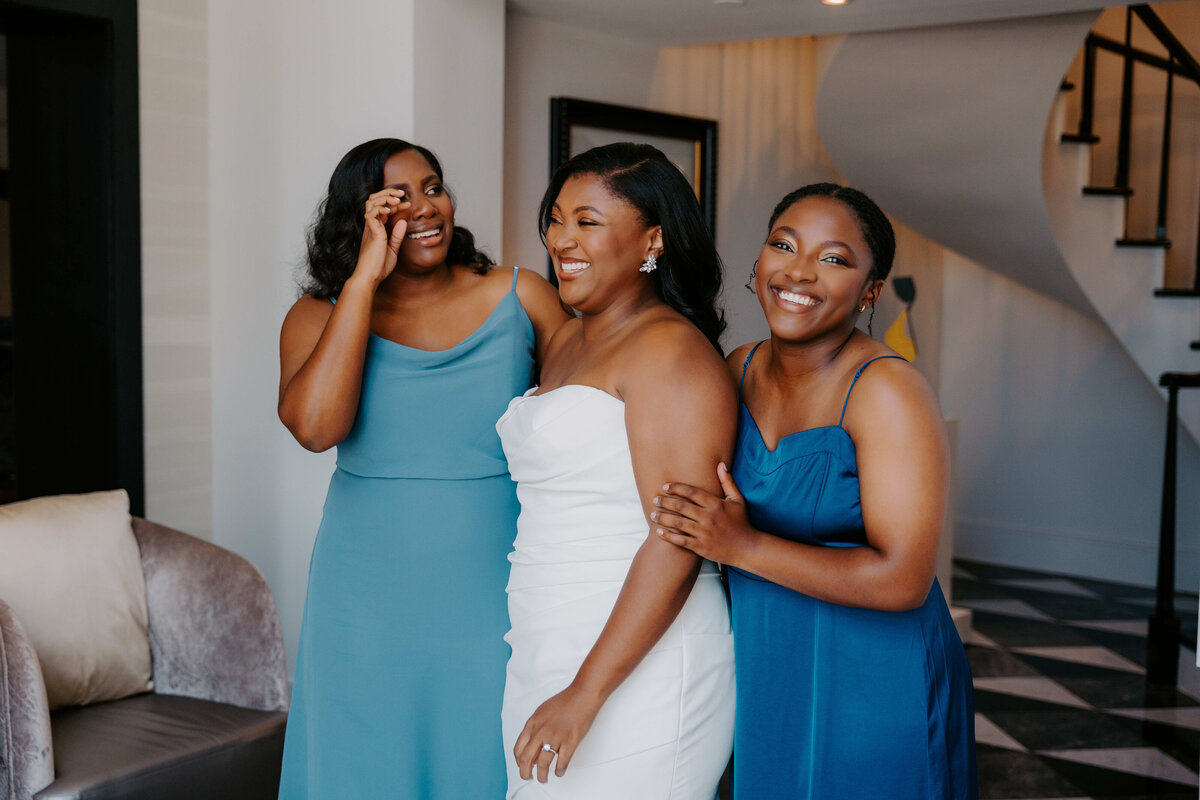 Three women, one in a white dress and two in teal dresses, smile joyfully in a modern room with a spiral staircase, conveying a celebratory mood.