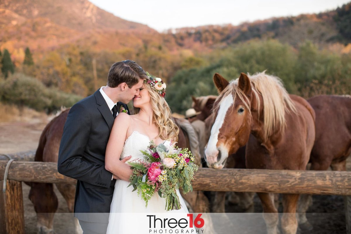 Bride looks back at her Groom and share a kiss while standing next to two horses at Riley's Farm