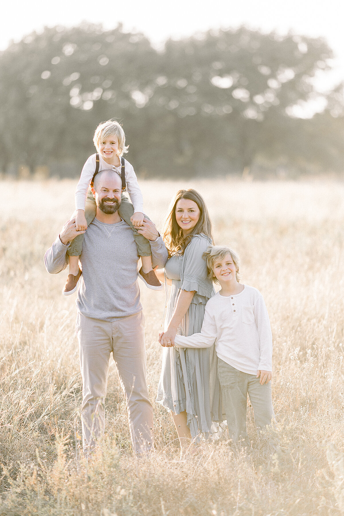 Beautiful family photo of a young family of 4 standing in the middle of an open grassy field at a Frisco park.