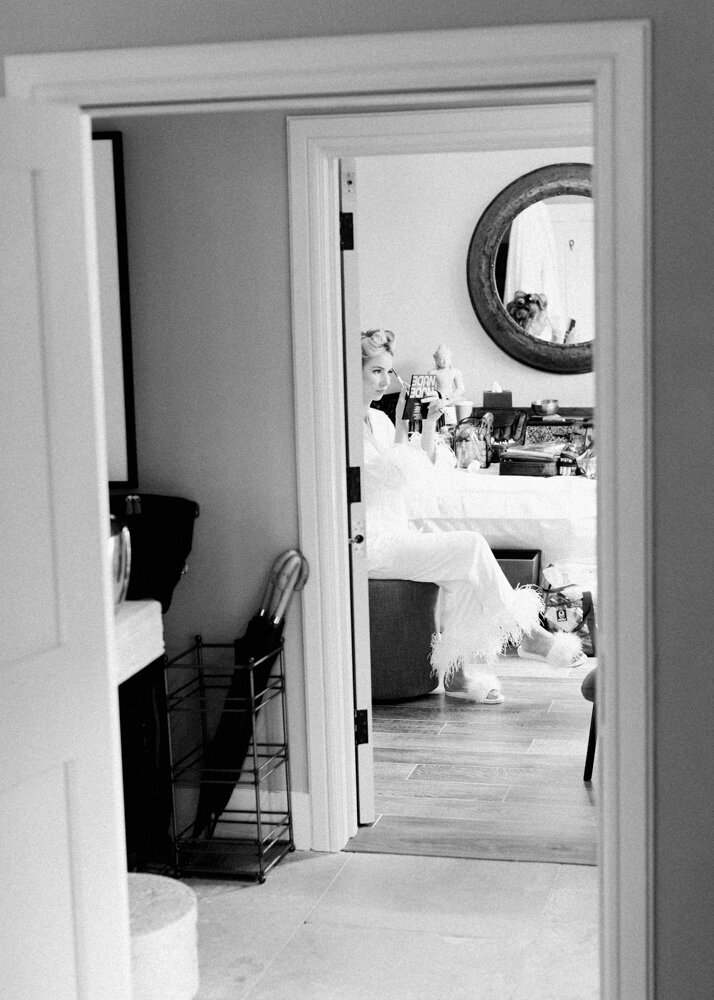 bride with hair rolls putting her makeup on while wearing white feather trim silk pyjama captured on black and white photo