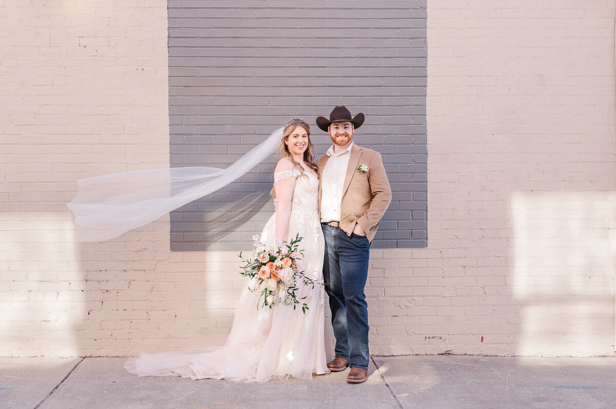 A smiling couple posing for portraits along side a white and grey brick wall with the bride's veil flying while enjoying their Raleigh wedding photography session with JoLynn Photography