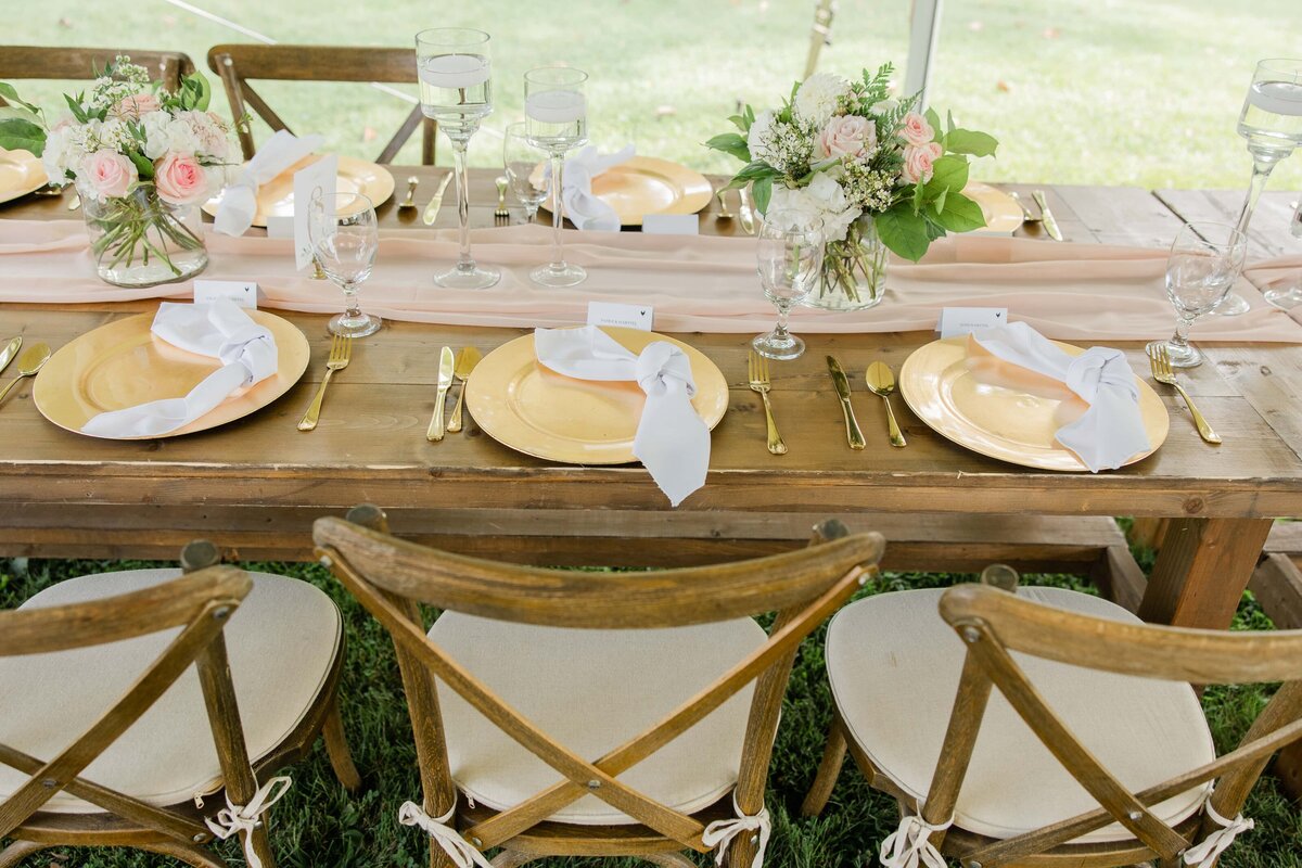 A wedding table setting at Park Farm Winery featuring golden plates, floral centerpieces, and wooden chairs with white ribbons under a tent.