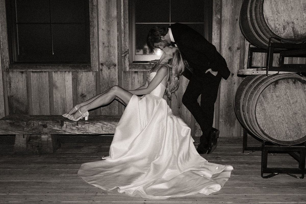A man in a suit kissing a woman in a flowing dress on a wooden bench, next to stacked barrels, in a rustic setting, possibly coordinated by a wedding planner from Des Moines.
