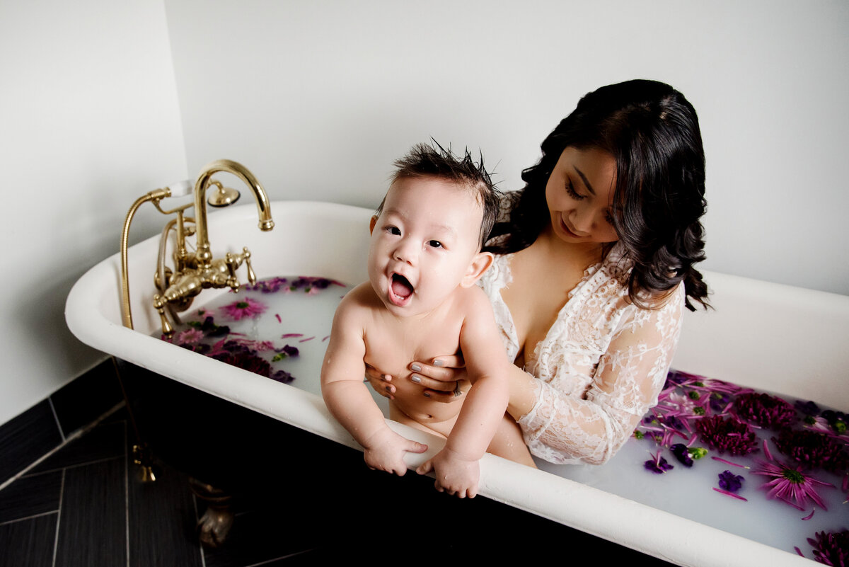 st-louis-motherhood-photographer-mother-holding-baby-in-milk-bath-with-purple-flowers-wearing-white-lace-gown