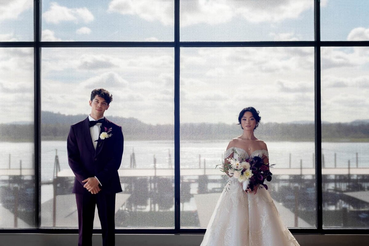 a stylish bride and groom stand in front of windows overlooking a marina of the columbia river
