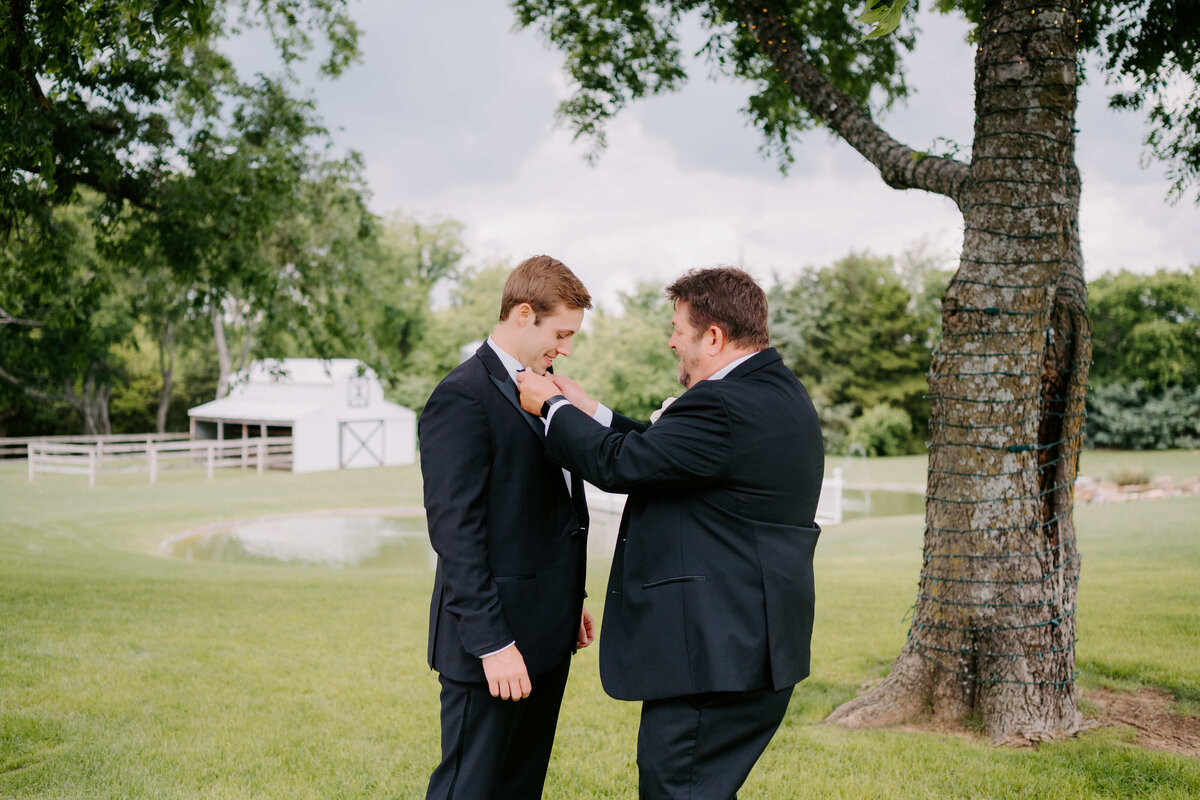 A man in a suit helps another adjust his tie outdoors by a tree. A barn and lush greenery are in the background, conveying a serene, supportive moment.