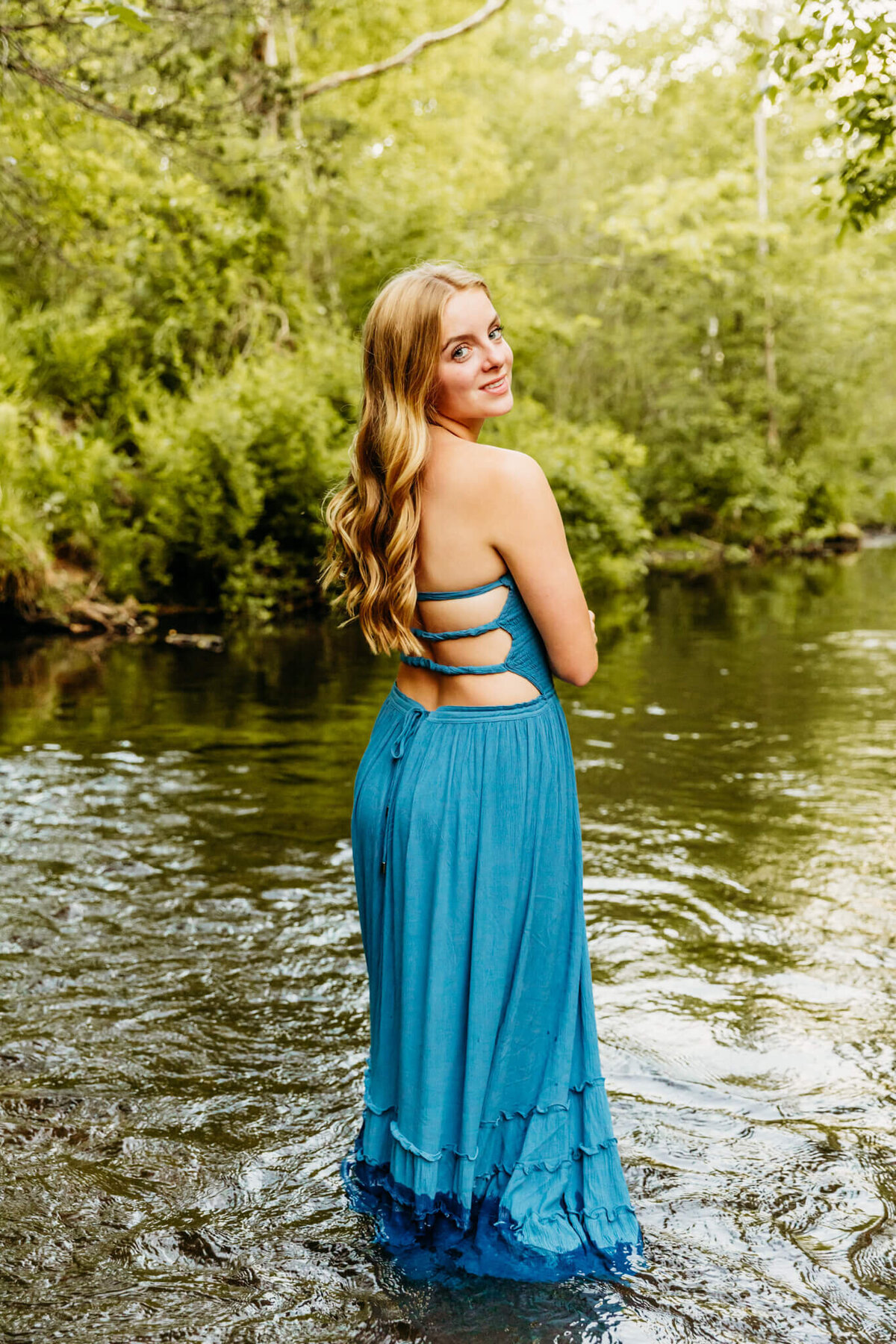 young lady in a long blue dress standing in a creek as she looks back over her shoulder captured by Green Bay Senior Photographer Ashley Kalbus