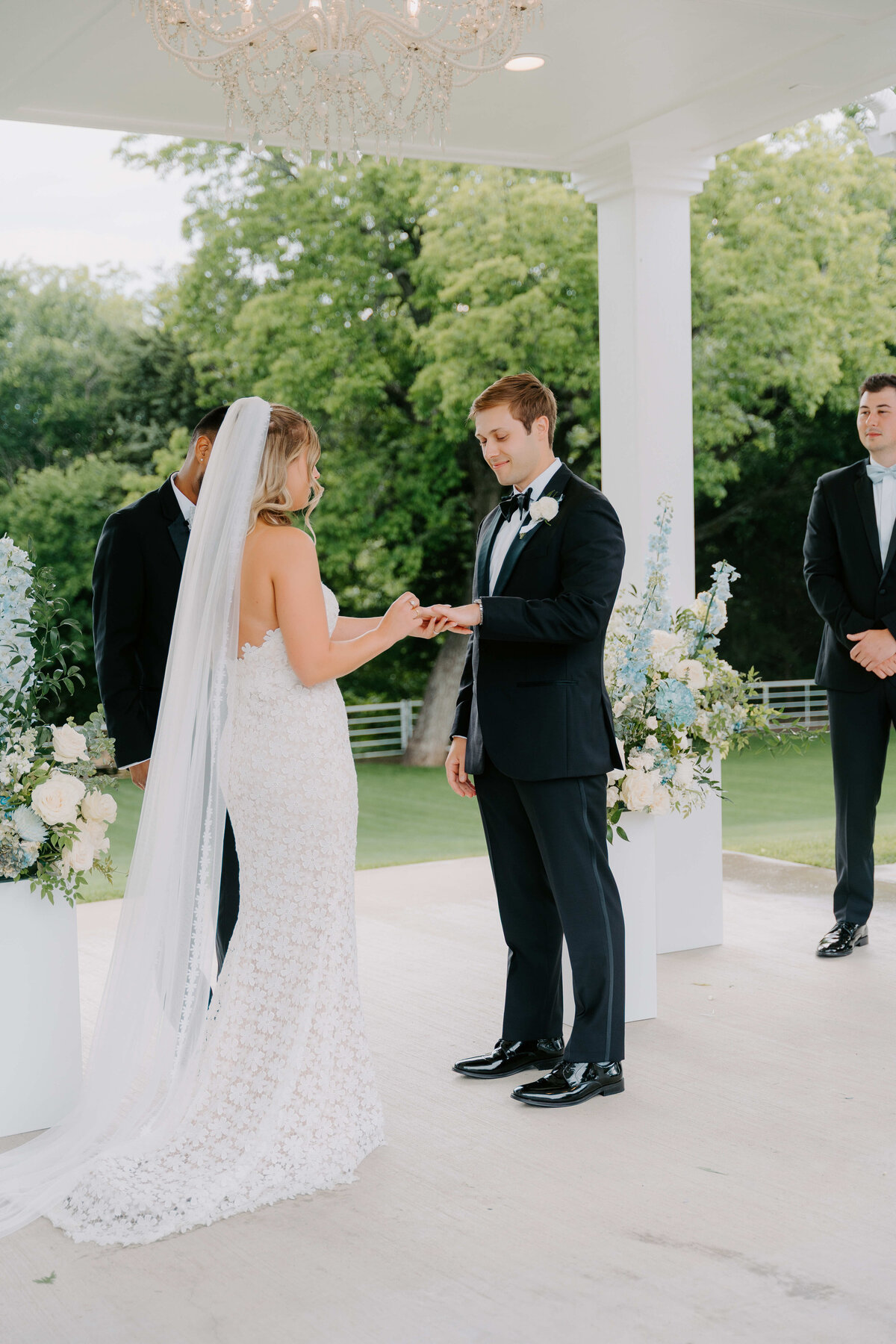 A bride and groom exchange rings under a white gazebo adorned with flowers. The scene is serene, with lush greenery in the background. The atmosphere is romantic and joyful.