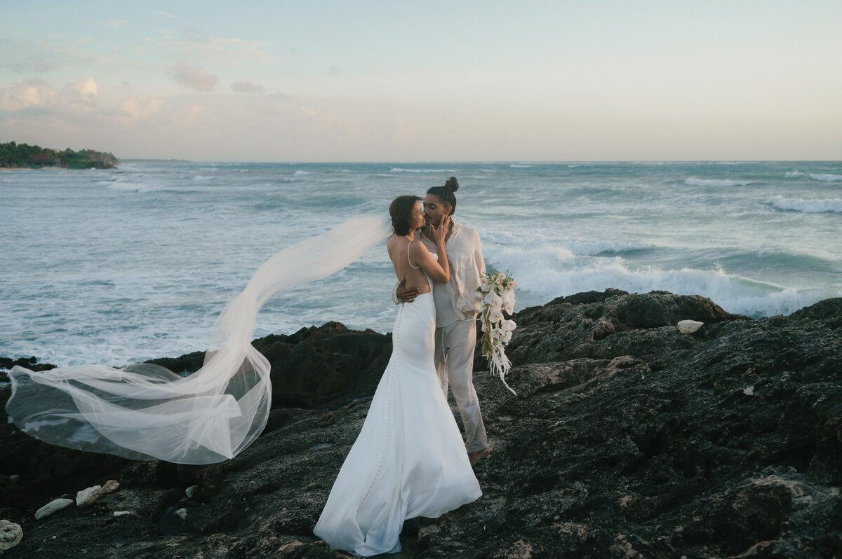 Wide angle of Greita and Jairo in wedding attire standing on a rocky cliff at Tulum Beach.