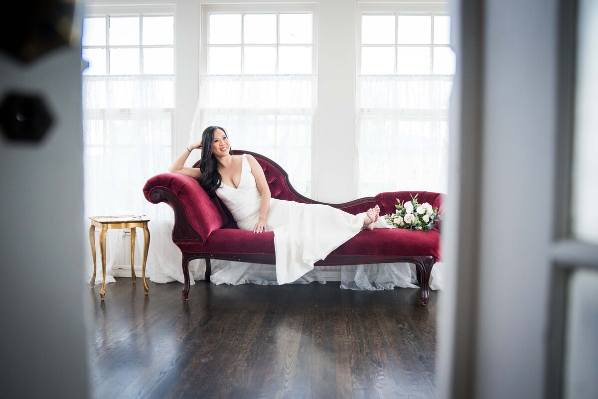 A bride poses with her bouquet on an elegant red velvet couch.