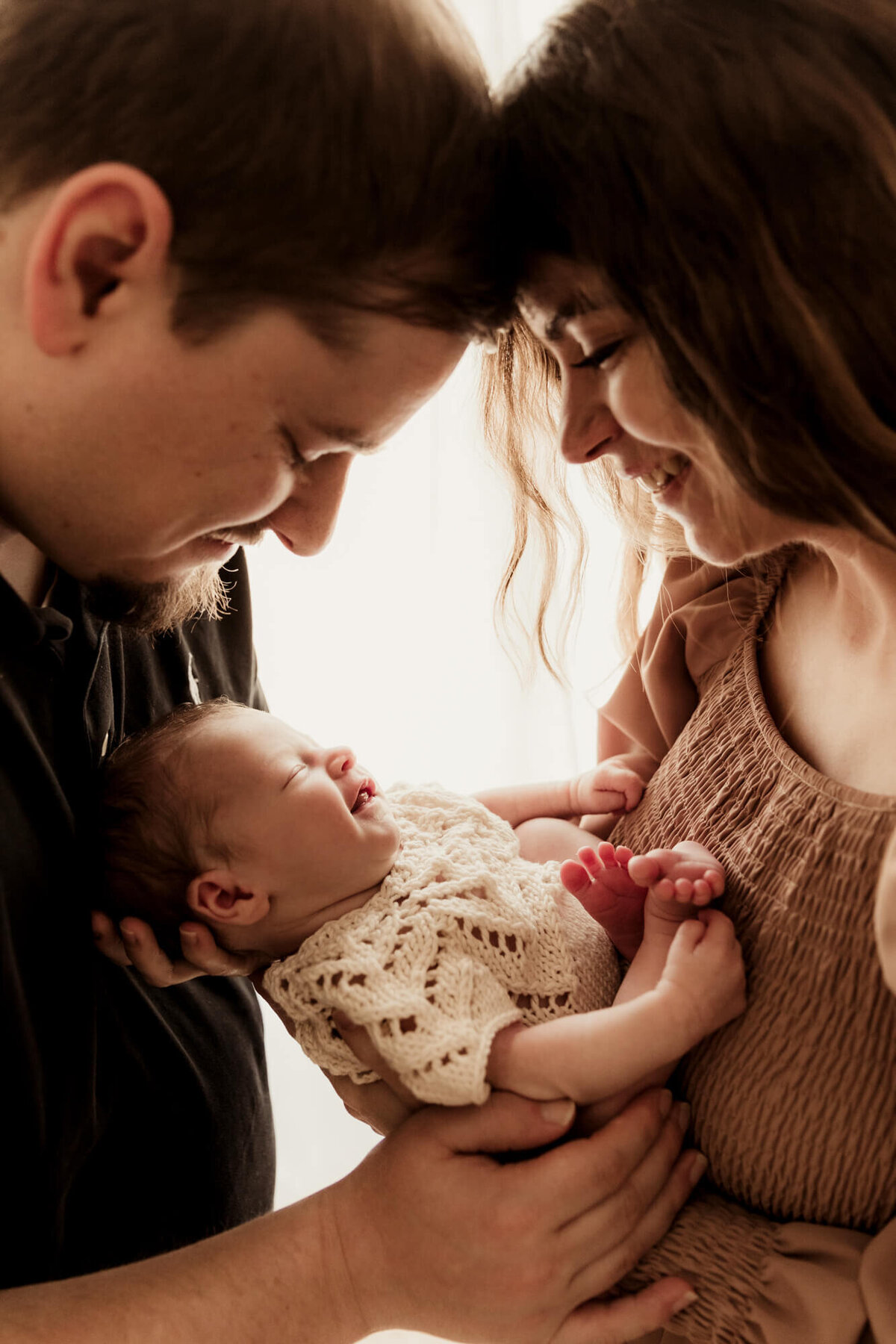 Baby girl smiles while mother and father hold her near a window for a newborn photo in OKC.