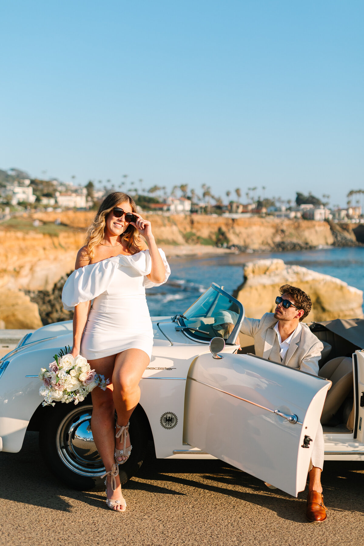 bride leaning against a vintage car while groom sits in driver's seat in front of a beach.