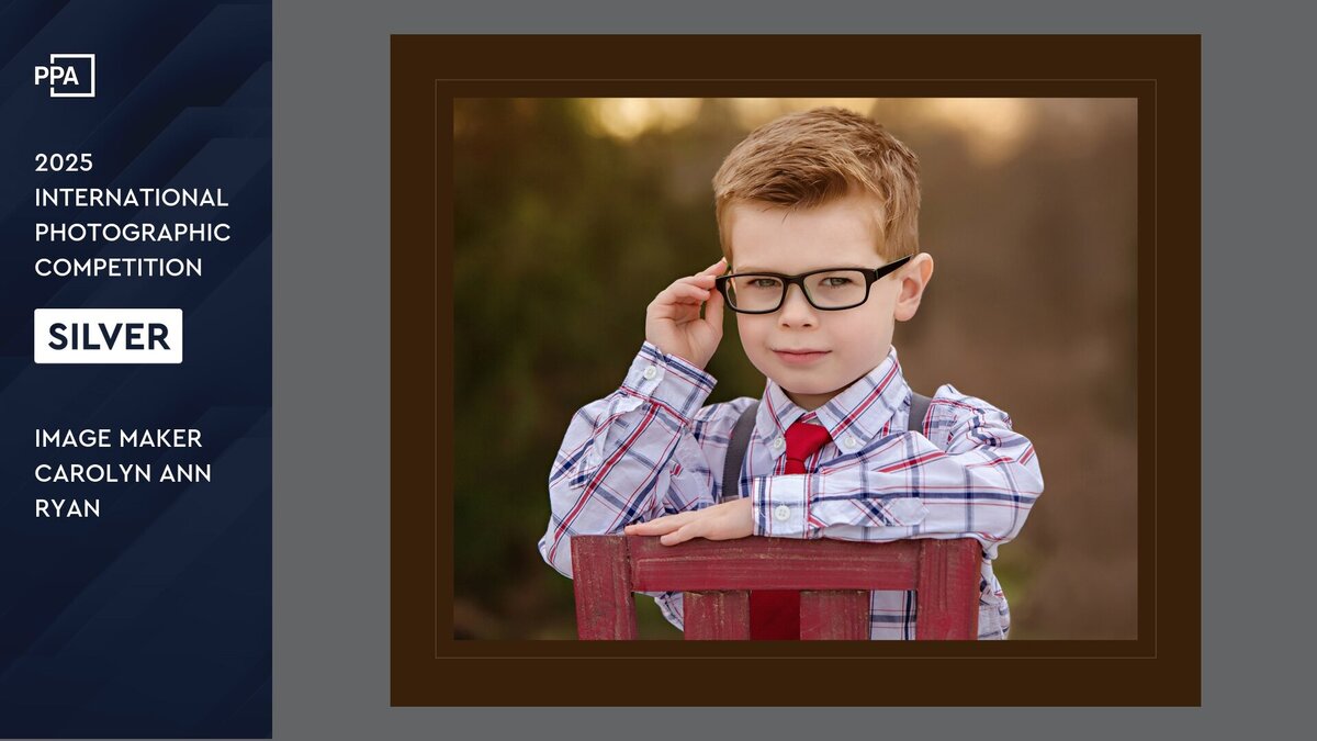 Photo of a young boy in a white and red plaid button down shirt and red tie sitting backwards on a red chair and one hand is touching his eyeglasses.  The side panel reads "PPA 2025 International Photographic Competition Silver Award Image Maker Carolyn Ann Ryan"