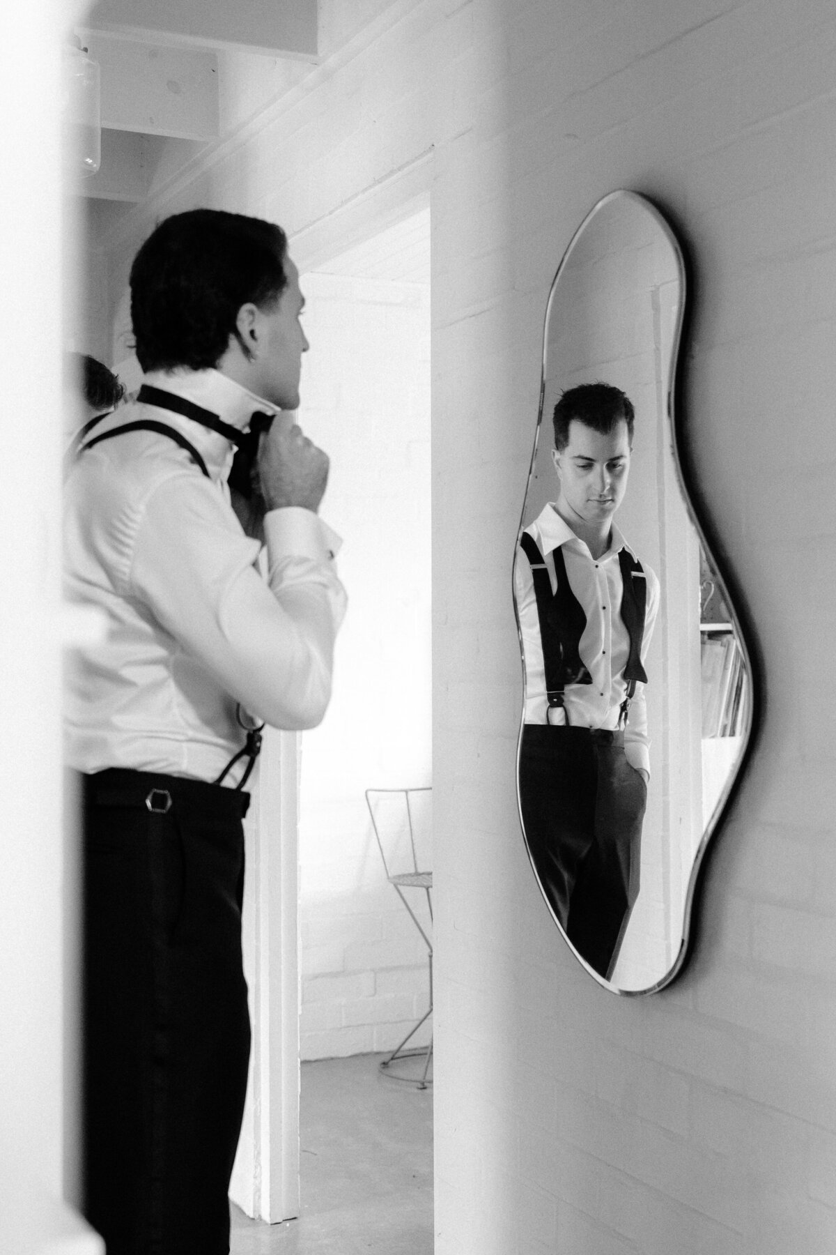 A groom wearing his bow tie in front of a mirror and his partner's reflection on the same mirror.