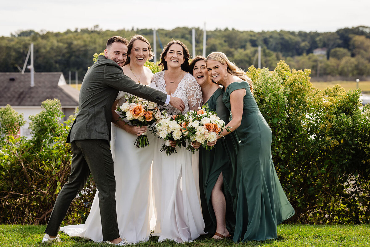 A group of two brides and three wedding party members, all smiling and posing for a photo outdoors, with the bridesmaids playfully pushing the brides together.