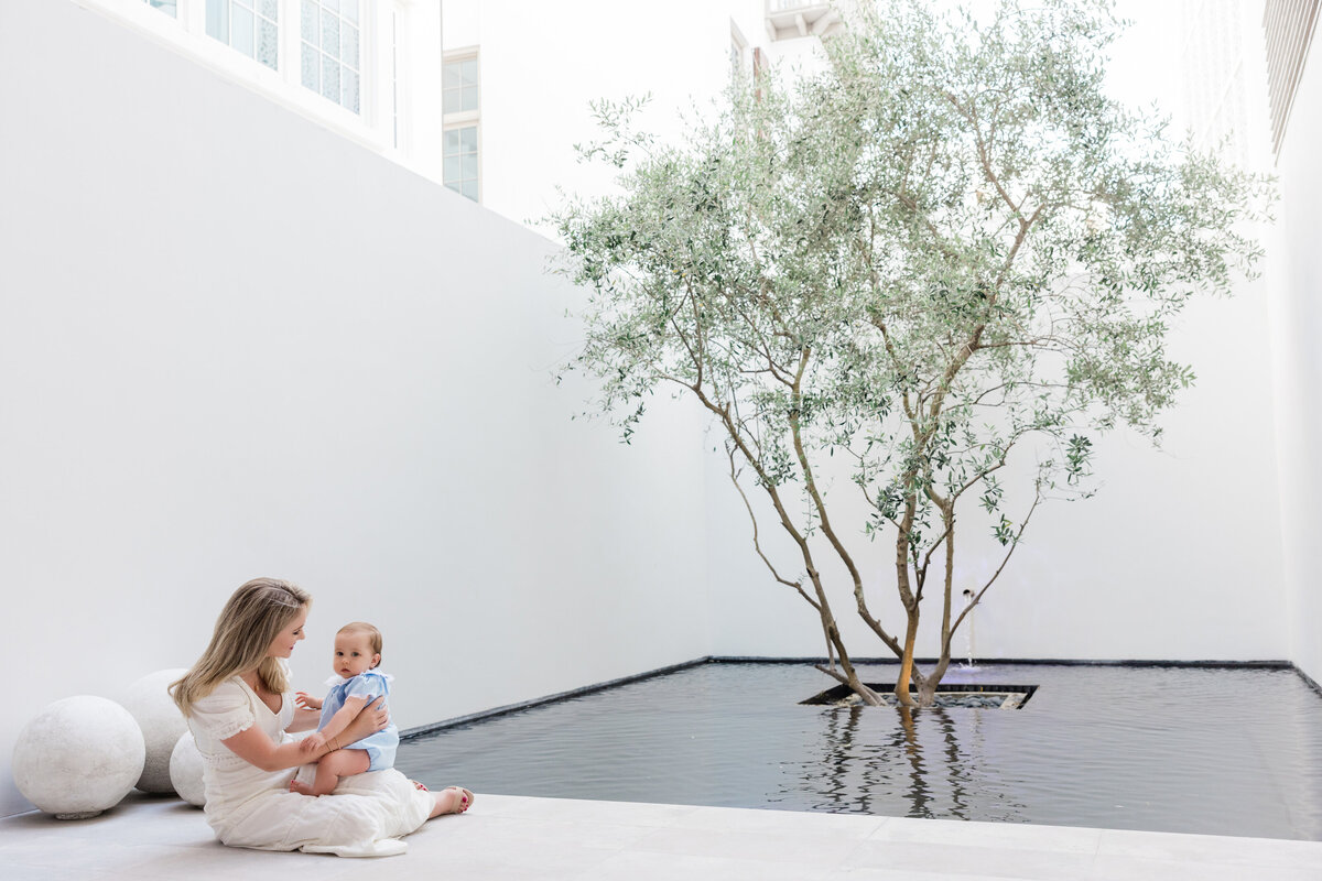 A woman sitting on the ground with her baby in her lap at the courtyard of a home in Alys Beach Florida. The photo is by a professional photographer.