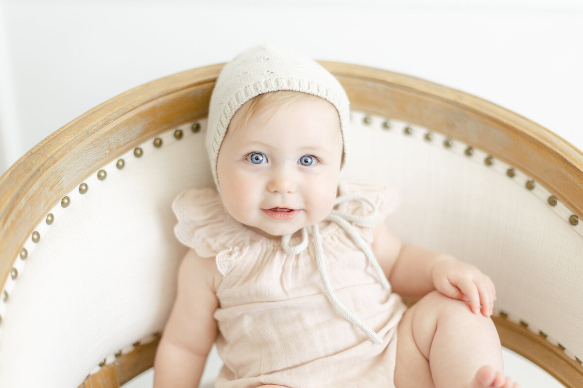 Cute baby girl with a knit bonnet on while she is sitting on a chair looking at the photographer in a DFW photography studio.