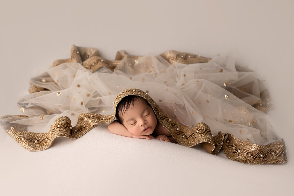 Fine art newborn photos captured by best Philadelphia newborn photographer Katie Marshall. Baby girl is sleeping on her belly with her hands folded underneath her chin. Top of her is draped cream organza fabric trimmed with gold silk and hold embellishments.