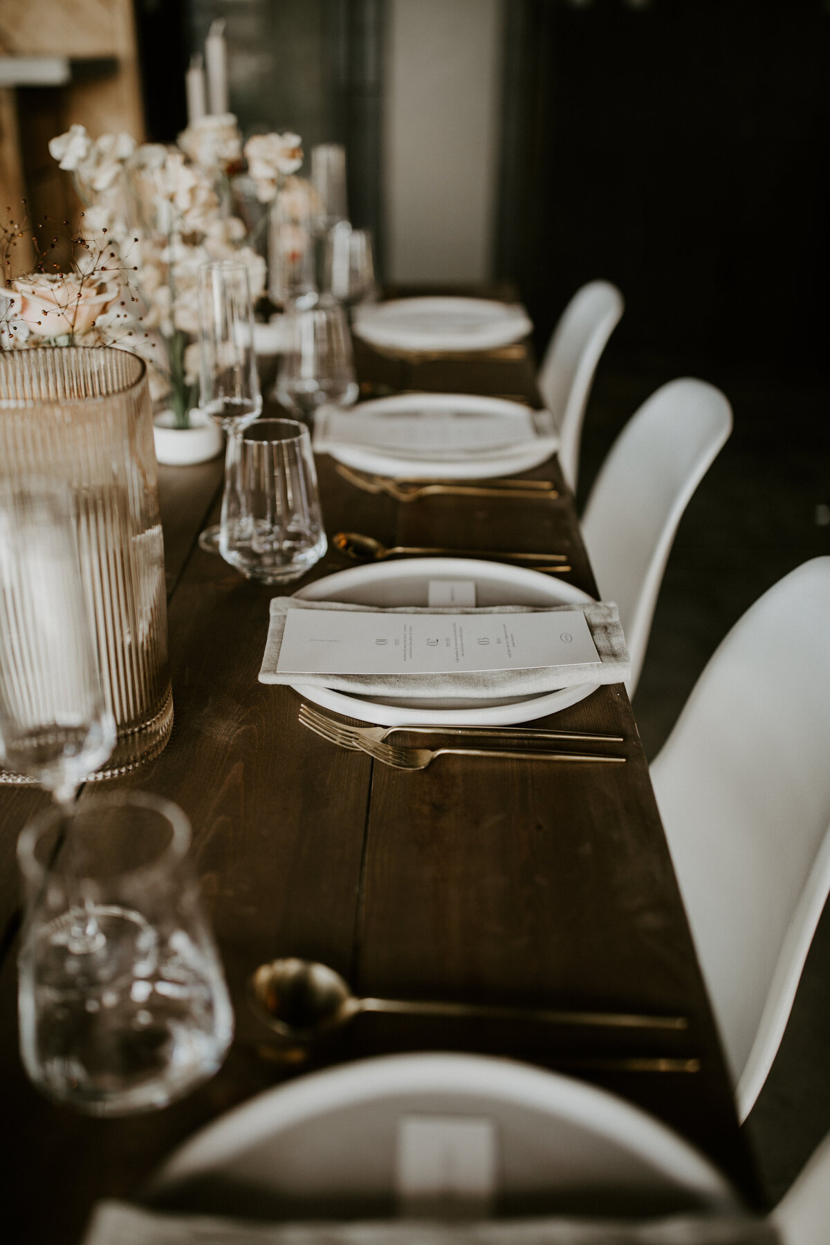 White dinner menus with black font atop a white linen napkin and white plate on brown wooden table with white chairs.