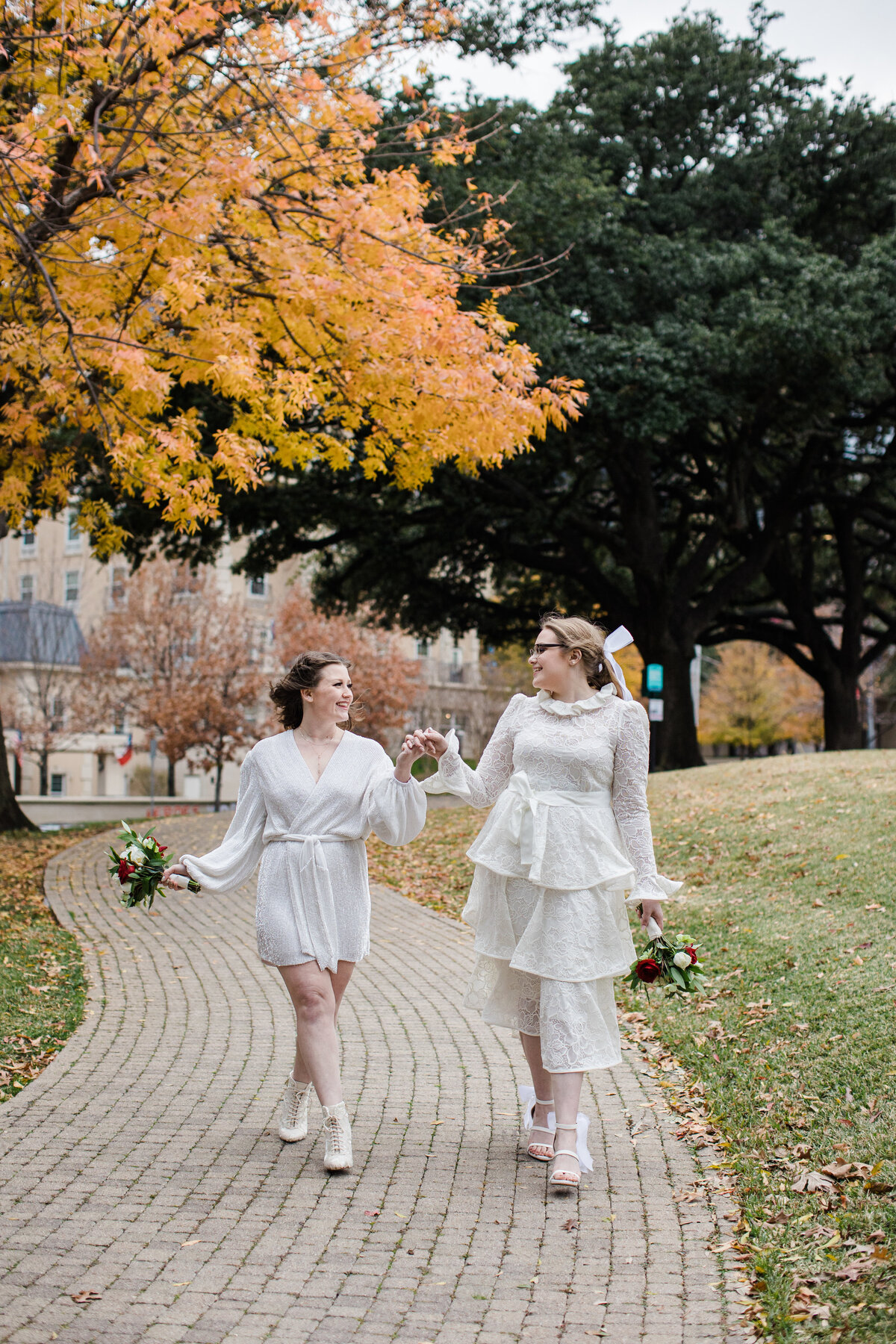 A portrait of two brides walking hand in hand down a path after their elopement ceremony at Turtle Creek in Dallas, Texas. The bride on the left is wearing a simple, elegant, short white dress while holding a bouquet. The bride on the right is wearing a layered, intricate, white dress also while holding a bouquet.