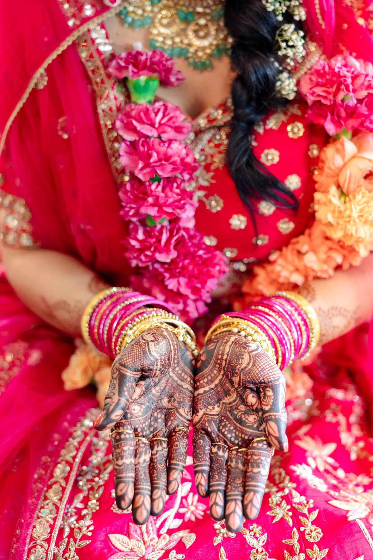A person wearing a vibrant red and gold embroidered outfit displays intricate henna designs on their hands. They are adorned with colorful bangles and floral garlands.