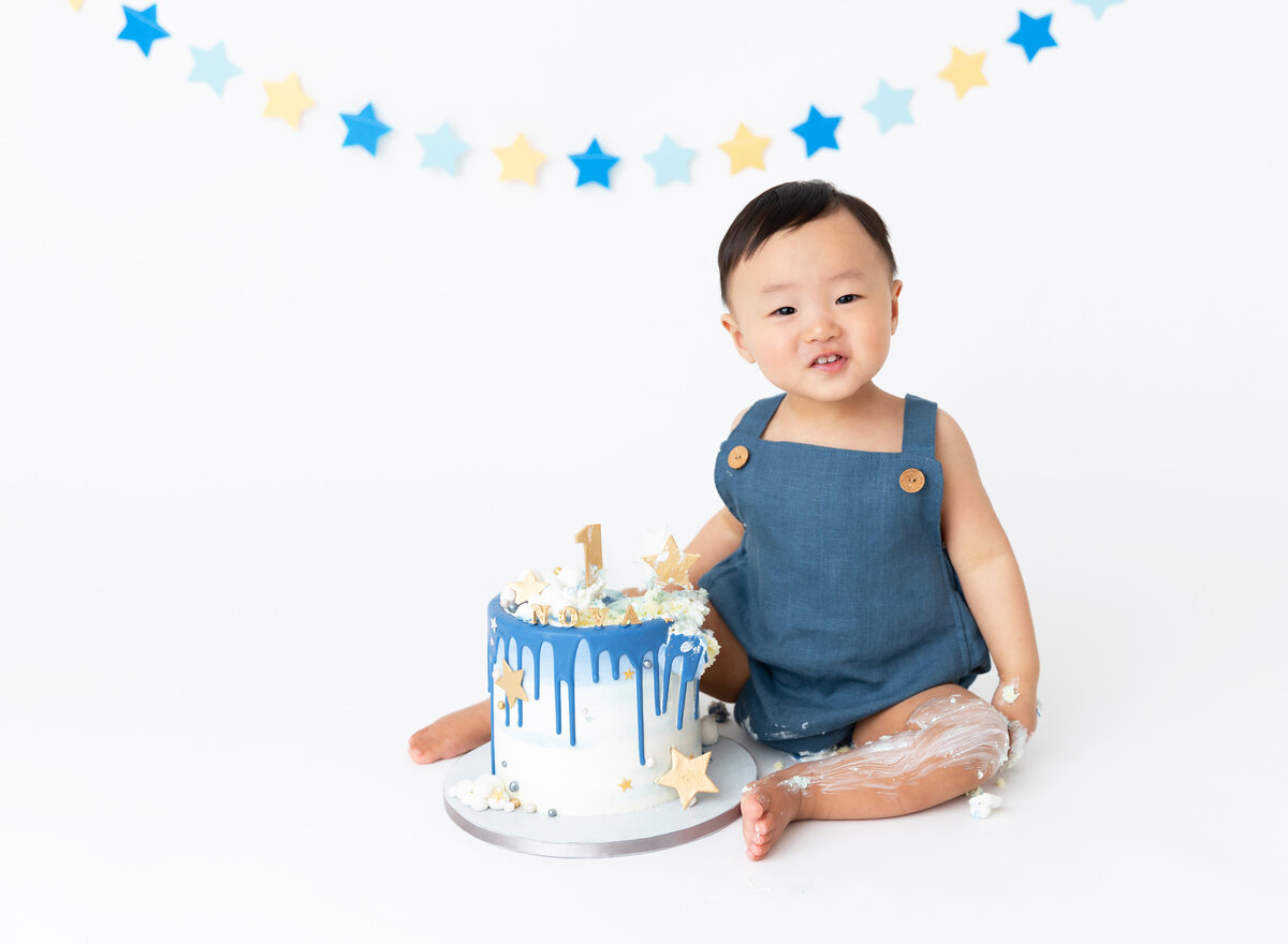 Baby boy waring a blue overall romper sits with icing on his hands and legs for first birthday cake smash photos.